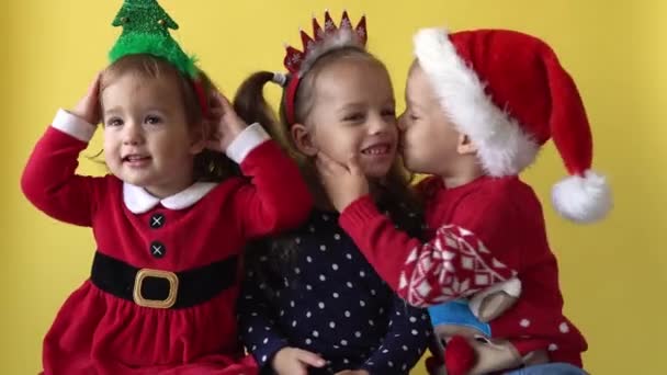 Émotion mignon heureux 3 frères et sœurs amis bébé fille et garçon baisers dans Santa costume regardant caméra à fond jaune. Jeu d'enfant Scène de Noël Célébration d'anniversaire. Enfant amusez-vous passer du temps Nouvel An — Video