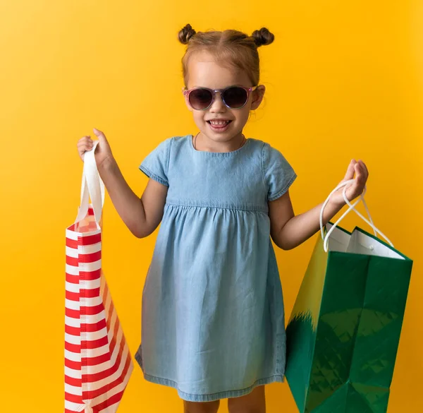 Retrato Hermosa niña feliz preescolar en gafas de sol sonriente alegre celebración bolsas de cartón aislado en el fondo de Orange Yellow Studio. Felicidad, Consumismo, Venta Gente comprando Concepto — Foto de Stock