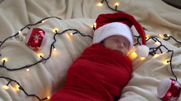 Top View Retrato Primeiros Dias de Vida Bebê Adormecido Engraçado Recém-Nascido Em Chapéu de Santa Envolvido Em Fralda Vermelha Em Fundo Garland Branco. Feliz Natal, Feliz Ano Novo, Bebê, Infância, Conceito de Inverno — Vídeo de Stock