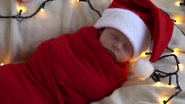 2015 년 12 월 9 일에 확인 함 . Top View Portrait First Days of Life Newborn Cute Funny Sleeping Baby In Santa Hat Wrapped In Red Diaper At White Garland Background. 메리 크리스마스, 해피 뉴 이어, 유아, 어린 시절, 겨울 컨셉 — 비디오