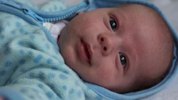 Kijk van de baby in de camera van dichtbij opgenomen. zuigeling, kindertijd, ouderlijke liefde, emotie concept - schattig lachend gezicht van bruin-ogige mollig pasgeborene in zacht blauw jasje wakker kijkt rond liggend op bed thuis — Stockvideo