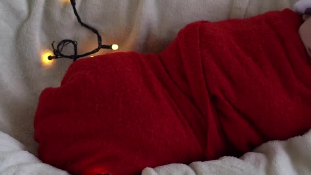 Yeni doğan bebeğin ilk portresi beyaz çelenk arka planında kırmızı beze sarılı, Noel Baba şapkalı komik uyuyan bebek. Mutlu Noeller, Mutlu yıllar, Bebek, Çocukluk, Kış Konsepti — Stok video