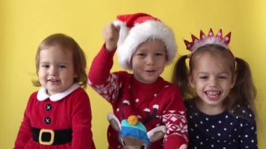 Duygusal Sevimli Mutlu Neşeli 3 Kardeş Arkadaş Bebek Kız ve Noel Baba Takım Elbiseli Çocuk sarı arka planda kameraya bakıyor. Doğum gününü kutlayan çocuk Noel Sahnesi oynuyor. Çocuk Yılbaşını Eğlenerek Geçiriyor