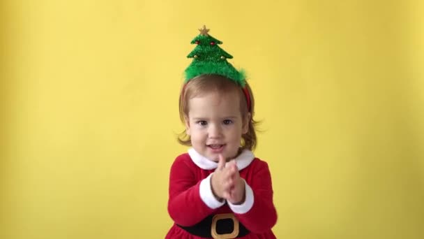 Porträtt Emotion Happy Chubby Toddler Baby Girl I Santa Suit leende applåder med julgran på huvudet tittar på kameran på gul bakgrund. Barn leker Fira. Ungen ha kul tillbringa nyår tid — Stockvideo