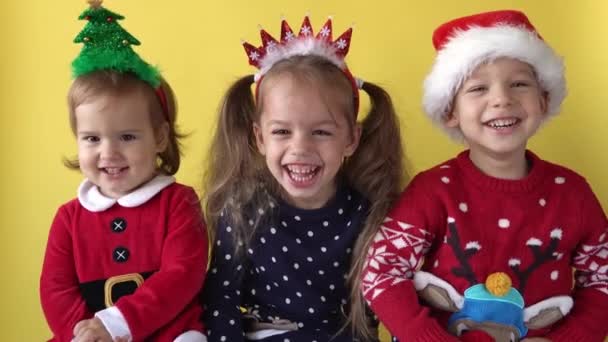 Эмоциональный милый Счастливые три сестры Друзья Девочка и Мальчик в костюме Санта-Клауса Смеясь глядя на камеру на желтом фоне. Дети празднуют Рождество. Детям весело провести Новый год — стоковое видео