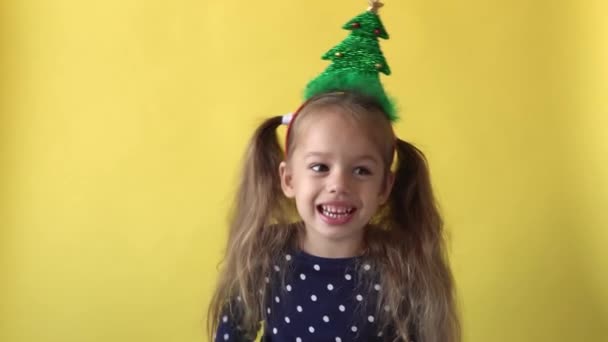 Retrato Emoción linda niña preescolar feliz con cola de caballo y árbol de Navidad en la cabeza cantando mirando a la cámara en el fondo amarillo. Niño celebrando el cumpleaños. El niño se divierte pasar el tiempo de año nuevo — Vídeo de stock