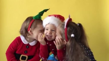 Duygusal Sevimli Mutlu 3 Kardeşler Kız Arkadaş, Öpüşen Bebek, Noel Baba Takımlı Kucaklaşma Sarı Arkaplan 'da kameraya bakıyor. Doğum gününü kutlayan çocuk Noel Sahnesi oynuyor. Çocuk Yılbaşını Eğlenerek Geçiriyor