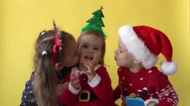 Эмоции милые Счастливые 3 Друзья Бэби Девочка и Мальчик Целуются в костюме Санта, глядя на камеру на желтом фоне. Детские игры Рождественская сцена празднования дня рождения. Детям весело провести Новый год — стоковое видео