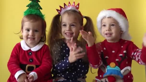 Συναίσθημα χαριτωμένο Happy 3 Αδέρφια Φίλοι κοριτσάκι και αγόρι applaud σε Santa κοστούμι κοιτάζοντας στην κάμερα σε κίτρινο φόντο. Παιδί Παίξτε Χριστουγεννιάτικη Σκηνή Γιορτάζοντας Γενέθλια. Καλά να περάσετε την Πρωτοχρονιά — Αρχείο Βίντεο