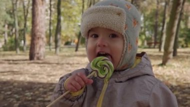 Ilık gri şapkalı küçük neşeli bebek çubukta büyük şeker yalıyor. Dışarıda şeker yiyen çocuk. Sarı Düşen Yapraklar Sonbahar Parkı 'nda Sepet Soğuk Hava' da. Çocukluk, Sonbahar