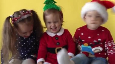 Duygusal şirin 3 kardeş, bebek kız arkadaş ve erkek çocuk Noel Baba kıyafetli ayak sallıyor ve sarı arka plandaki kameraya bakıyor. Doğum gününü kutlayan çocuk Noel Sahnesi oynuyor. Çocuk Yılbaşını Eğlenerek Geçiriyor