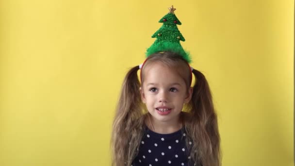 Πορτρέτο Συναίσθημα χαριτωμένο χαρούμενο κορίτσι προσχολικής ηλικίας με αλογοουρά και χριστουγεννιάτικο δέντρο στο κεφάλι χαμογελώντας κοιτάζοντας στην κάμερα σε κίτρινο φόντο. Παιδί γιορτάζει γενέθλια. Καλά να περάσετε την Πρωτοχρονιά — Αρχείο Βίντεο