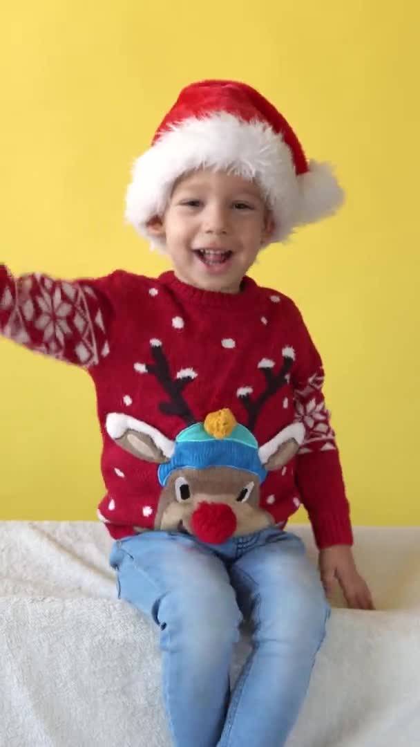 垂直的肖像情感可爱快乐有趣的学龄前男婴在圣诞老人的帽子里嬉笑着看相机。黄色背景。儿童圣诞庆典。「小朋友过年快乐」 — 图库视频影像