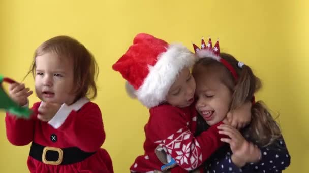 Συναίσθημα χαριτωμένο Happy 3 Αδέρφια Φίλοι Μωρό Αγόρι Κορίτσι Φιλί Αγκαλιά Σε Σάντα κοστούμι κοιτάζοντας στην κάμερα σε κίτρινο φόντο. Παιδί Παίξτε Χριστουγεννιάτικη Σκηνή Γιορτάζοντας Γενέθλια. Καλά να περάσετε την Πρωτοχρονιά — Αρχείο Βίντεο