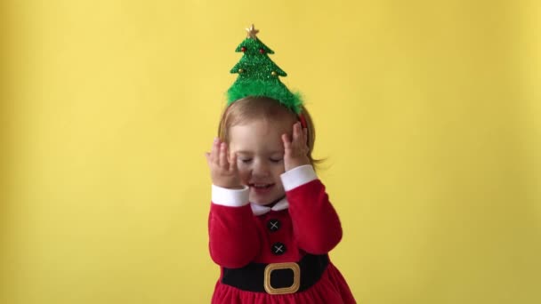 Retrato Emoción Feliz Niño Regordeta Bebé en traje de Santa. Niño sonriendo lindo cierra los ojos con las manos Árbol de Navidad en la cabeza mirando a la cámara en el fondo amarillo. El niño se divierte pasar el tiempo de año nuevo — Vídeos de Stock