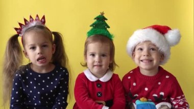 Duygusal Sevimli Mutlu Neşeli 3 Kardeş Arkadaş Bebek Kız ve Noel Baba Takım Elbiseli Çocuk sarı arka planda kameraya bakıyor. Doğum gününü kutlayan çocuk Noel Sahnesi oynuyor. Çocuk Yılbaşını Eğlenerek Geçiriyor
