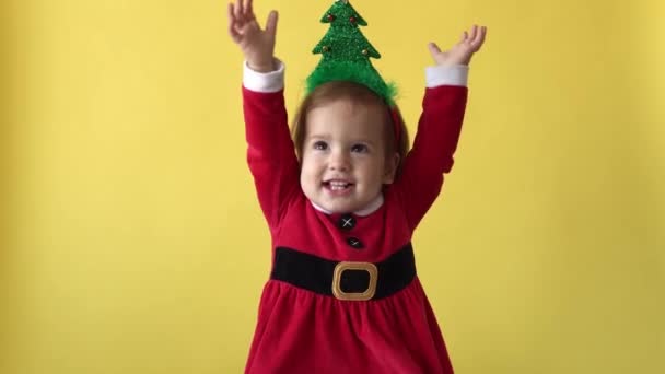 Retrato emoção feliz gordinha criança bebê menina em Santa terno levanta as mãos com árvore de Natal na cabeça olhando para a câmera no fundo amarelo. Criança Brincar Celebre. Criança Divirta-se Passe o Ano Novo — Vídeo de Stock