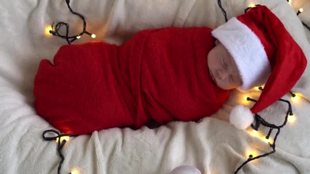 Top View Portret Primele zile de viață Nou-născut drăguț amuzant adormit copil în pălărie Moș Crăciun înfășurat în scutec roșu la fundal Garland alb. Crăciun fericit, An Nou fericit, Copil, Copilărie, Concept de iarnă — Videoclip de stoc