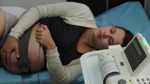 Молодая беременная женщина на больничном диване во время кардиотокографии. Посетите врача-гинеколога в клинике для беременных консультантов. Проверка на беременность беременной матери живота — стоковое видео