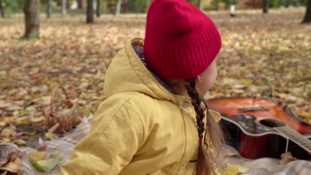 Close up kleine vrolijke kleuter in warme rode hoed eet likken grote snoep op stok. Kind eten snoep buiten.Gele Gevallen bladeren In Mand Koud Weer In Fall Park. Jeugd, herfst — Stockvideo