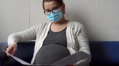 Genç Hamile Koruyucu Maskeli Kadın, hastane için randevu bekleyen koltukta kitap okuyor. Klinikteki jinekoloğu ziyaret et. Muayene Edilmeyi Bekleyen Göbek Sağlığı Kontrolü