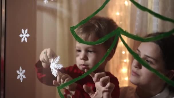 Auténtica mamá linda y dos niños pequeños preescolares hermanos niña y niño de 2-4 años dibujan el árbol de Navidad en la ventana. mujer joven con niños pega copos de nieve. Navidad, Año Nuevo, concepto de invierno. — Vídeo de stock