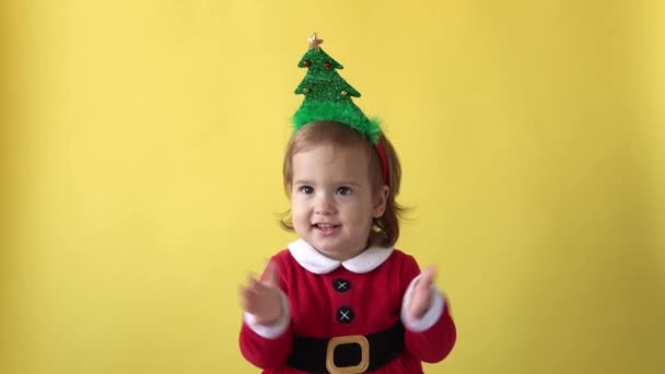 Retrato emoção feliz gordinha criança bebê menina em Santa terno sorrindo Applaud com árvore de Natal na cabeça olhando para a câmera no fundo amarelo. Criança Brincar Celebre. Criança Divirta-se Passe o Ano Novo — Vídeo de Stock