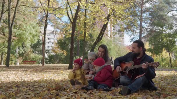 Duża szczęśliwa rodzina z małymi dziećmi relaksującymi się na pikniku spędzają razem czas, tata gra na gitarze śpiewając śmieszne piosenki. Spójrz na kamerę Zimna pogoda w jesiennym parku. Dzieciństwo, Rodzina, Jesienna koncepcja — Wideo stockowe