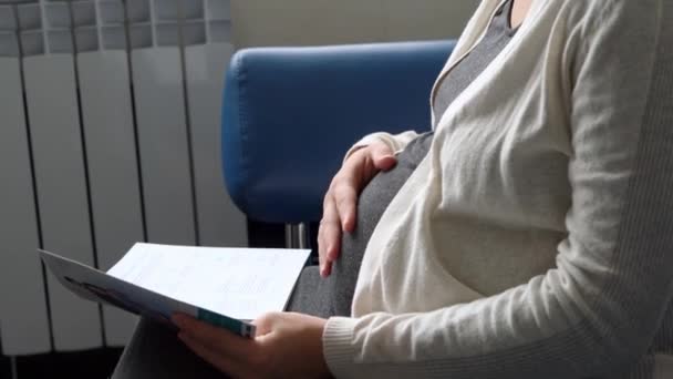 Молода вагітна жінка в медицині Захисна маска Читання на дивані чекає призначення медичного контролю над лікарнею. Відвідайте гінеколога доктора в клініці. Перевірка очікуваного здоров'я живота Перевірте — стокове відео