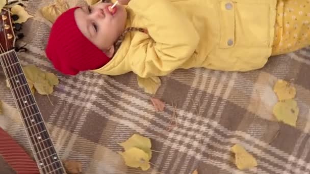 Arriba ver Pequeña niña preescolar Sonriente tumbarse descansando relajarse En cuadros hojas caídas En la cesta Picnic Niños lame Comer dulces caramelos Mira la cámara en Fall Park. Familia, Concepto de otoño — Vídeos de Stock