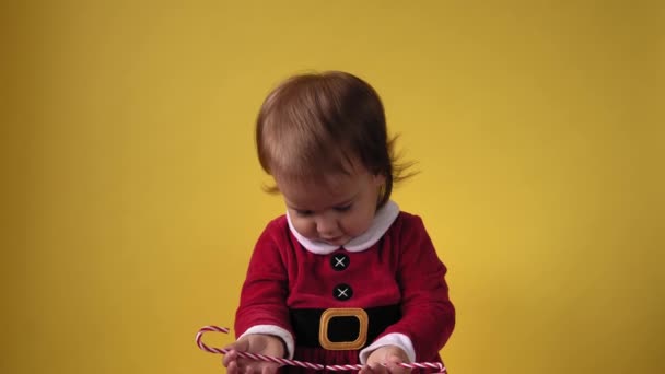 Cute Happy Wesoły Chubby Maluch dziewczynka w Santa Garnitur patrząc na aparat fotograficzny na żółtym tle. Dziecko bawiące się na scenie z okazji urodzin. Kid smak jedzenia lizać Boże Narodzenie słodkie cukierki pałeczki Nowy Rok — Wideo stockowe