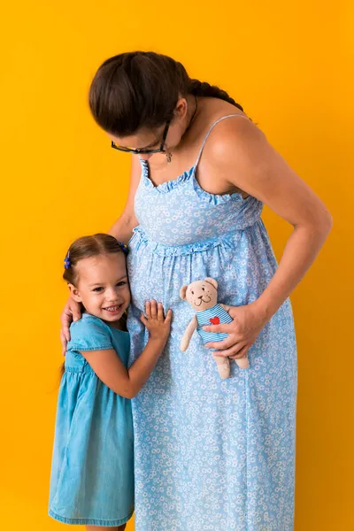 Материнство, любовь, детство, беременность, жаркое лето - обрезанный портрет беременной неузнаваемой матери женщины голубое платье маленькая дочка с братом и сестрой Тедди медвежонок обнимает маму животом на желтый — стоковое фото