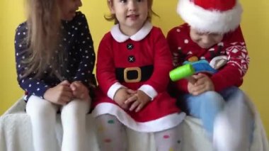 Duygusal şirin 3 kardeş, bebek kız arkadaş ve erkek çocuk Noel Baba kıyafetli ayak sallıyor ve sarı arka plandaki kameraya bakıyor. Doğum gününü kutlayan çocuk Noel Sahnesi oynuyor. Çocuk Yılbaşını Eğlenerek Geçiriyor