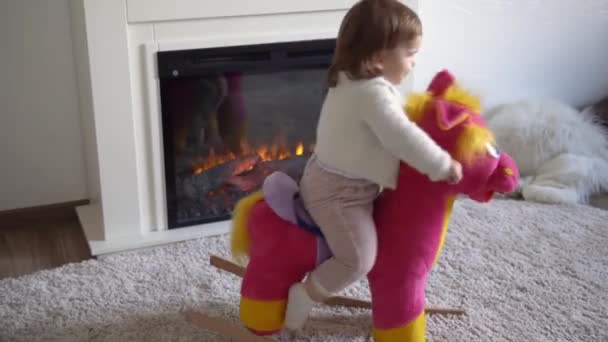 Familie jeugd. Happy kleine peuter kind meisje warme trui winter kleding spelen uitkleden op zacht paard in de kamer in de buurt van open haard. Leuke speelse authentieke Kid speelt thuis alleen swing op Toy — Stockvideo