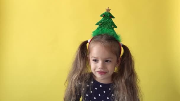 Πορτρέτο Συναίσθημα χαριτωμένο χαρούμενο κορίτσι προσχολικής ηλικίας με αλογοουρά και χριστουγεννιάτικο δέντρο στο κεφάλι χαμογελώντας κοιτάζοντας στην κάμερα σε κίτρινο φόντο. Παιδί γιορτάζει γενέθλια. Καλά να περάσετε την Πρωτοχρονιά — Αρχείο Βίντεο