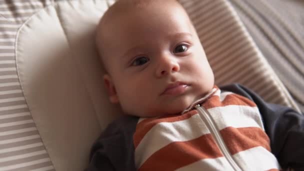 Ματιά του μωρού στην κάμερα κοντά στο βρέφος, παιδική ηλικία, γονική αγάπη. Χαριτωμένο χαμογελαστό νεογέννητο πρόσωπο σε ριγέ πιτζάμες σώματος ξύπνιοι ματιά γύρω για την απαλή καρέκλα στο σπίτι. χαρούμενη ζωή του ευτυχισμένου παιδιού — Αρχείο Βίντεο
