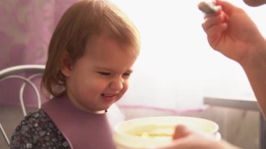Yemek, beslenme, çocukluk kavramları. Anne bebeğini kaşık kaşık ile besliyor. Önlük giymiş şirin, aç bir çocuk çocuk çocuk sandalyesine oturur. Kapalı tabaktan yemek yemek istemez. Önce sevme