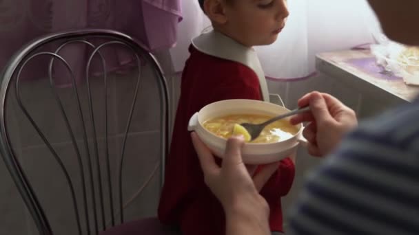 食物、营养、童年时期的概念- -母亲用橡胶勺子喂小孩。穿着围裙的饥饿可爱的孩子坐在孩子们的椅子上，不想在室内用盘子吃饭。不喜欢第一 — 图库视频影像