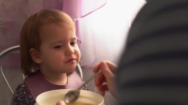 Yemek, beslenme, çocukluk kavramları. Anne bebeğini kaşık kaşık ile besliyor. Önlük giymiş şirin, aç bir çocuk çocuk çocuk sandalyesine oturur. Kapalı tabaktan yemek yemek istemez. Önce sevme