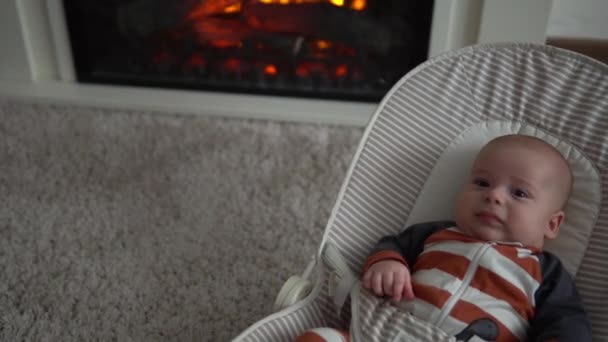 看着婴儿在相机里的婴儿，童年，父母的爱。可爱的笑脸，穿着条纹紧身衣的新生儿醒着躺在壁炉边的椅子上，在家里。快乐的生活幸福的孩子 — 图库视频影像
