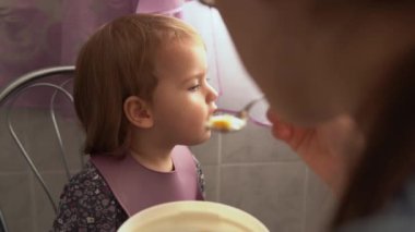 Yemek, beslenme, çocukluk kavramları. Anne bebeğini kaşık kaşık ile besliyor. Önlük giymiş şirin, aç bir çocuk çocuk çocuk sandalyesine oturur. Kapalı tabaktan yemek yemek istemez. Önce sevme
