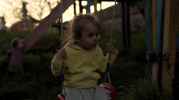 Authentique jolie petite fille d'âge préscolaire en gris jaune monte sur la vieille balançoire dans le parc sur l'herbe au coucher du soleil de printemps. enfant sur la nature au lever du soleil. Enfance, parentalité, famille, mode de vie concept — Video