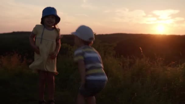 Schattig gelukkig kleuter baby paar knuffelen springen en rommelen rond op de top van de berg in hoog gras voor zonsondergang. Kind wandelen in het veld weide. kindertijd, natuur, levensstijl, vriendschap, zomerconcept — Stockvideo