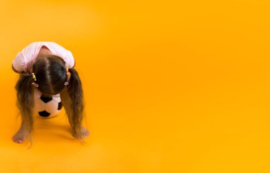 Klasik siyah-beyaz futbol topuyla gülümseyen sevimli anaokulu kızı sarı arka plandaki kameraya bakıyor. Çocuk tişört ve şortla futbol oynuyor. Spor, şampiyonluk, takım konsepti.