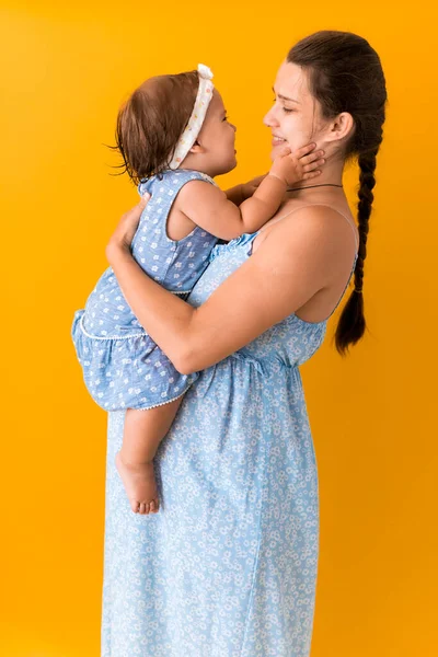 Материнство, жаркое лето - портрет молодой беременной счастливой улыбающейся матери женщины в синем платье, держа маленькую дочурку малыша, брата, босых ног в руках, глядя в глаза поцелуй на желтом фоне — стоковое фото