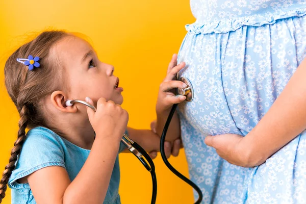 Motherhood, hot summer - croped portrait pregnant mother unrecognizable woman blue dress little Daughter girl sibling treat mom role play game stethoscope unborn big belly tummy on yellow background — Stock Photo, Image