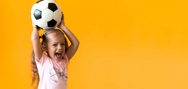 Hiteles aranyos mosolygós óvodás kislány klasszikus fekete-fehér focilabda nézni kamera sárga háttérrel. A gyerek pólóban és rövidnadrágban focizik. Sport, bajnokság, csapatkoncepció Stock Fotó