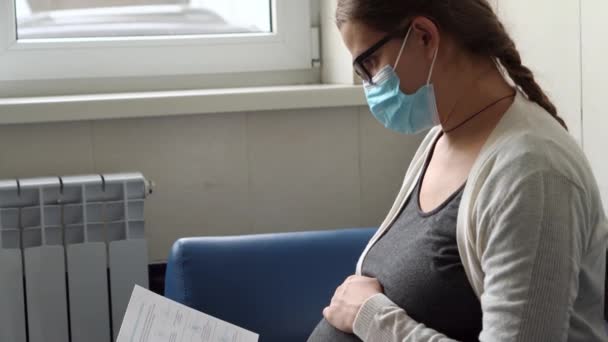Молода вагітна жінка в медицині Захисна маска Читання на дивані чекає призначення медичного контролю над лікарнею. Відвідайте гінеколога доктора в клініці. Перевірка очікуваного здоров'я живота Перевірте — стокове відео