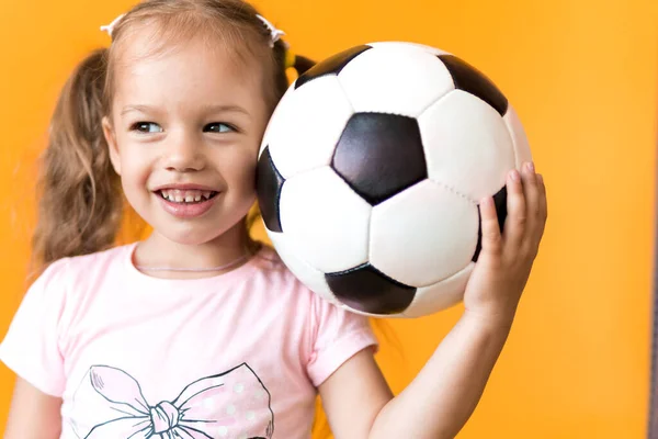 Hiteles aranyos mosolygós óvodás kislány klasszikus fekete-fehér focilabda nézni kamera sárga háttérrel. A gyerek pólóban és rövidnadrágban focizik. Sport, bajnokság, csapatkoncepció Stock Kép