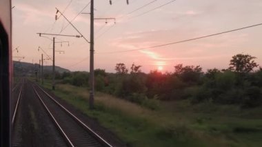 Pencereden yüksek hızlı tren manzarası güzel doğa manzarası vahşi arazi ve orman rayları yaz arkaplanında akşam günbatımında tren rayları. Ulaşım, seyahat, demiryolu, iletişim konsepti